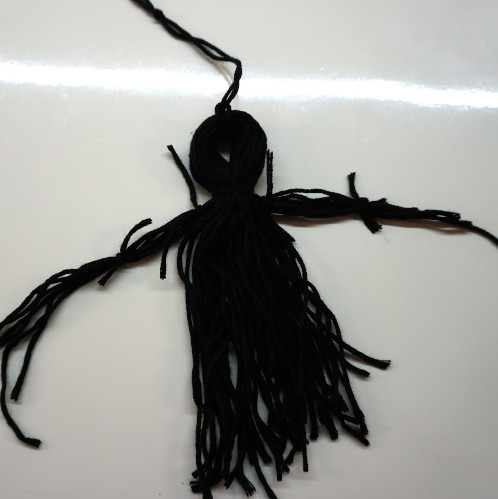 Die schwarze Zauberer-Figur hat schon einen Kopf und Hals. Es werden Wollfäden für die Arme und Beine abgebunden