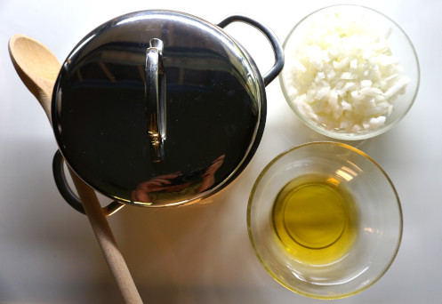 Topf mit geschlossenem Deckel, daneben ein Kochlöffel, ein Schälchen mit Öl, ein Schälchen mit geschnittenen Zwiebeln